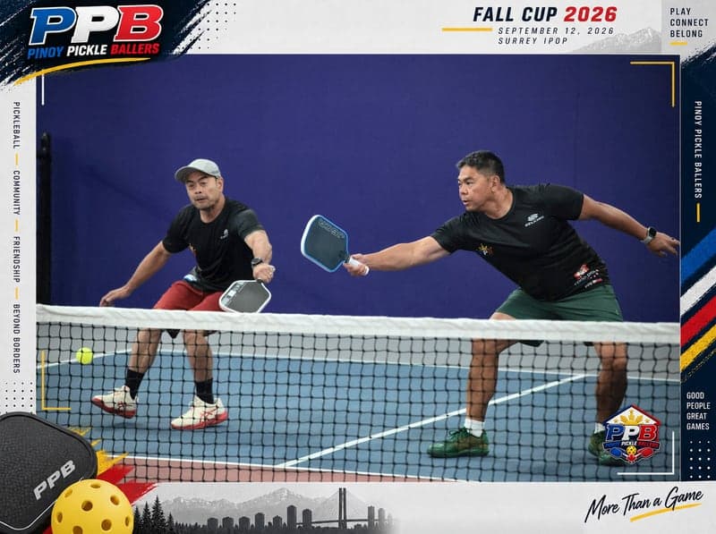Doubles partners stretch toward a low ball at the net on an indoor court with a purple wall behind them.